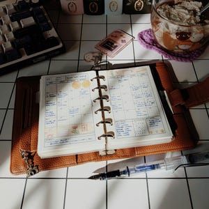 May include: An open brown leather ring-bound planner with a monthly calendar grid filled with handwritten notes, a clear fountain pen, a small pink keychain, and a keyboard on a white grid surface.