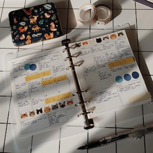 Pode incluir: Uma agenda de seis argolas aberta com papel quadriculado, notas manuscritas e autocolantes de gatos. Ao lado, uma lata retangular com padrão de animais da floresta, dois rolos de fita adesiva e uma caneta de tinta permanente sobre uma grelha.