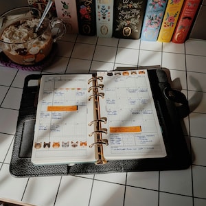 May include: A black leather six-ring planner open to a monthly calendar page with cat-themed stickers, handwritten notes, and orange sticky tabs. The planner rests on a white grid surface near a glass mug of coffee and several colorful books.