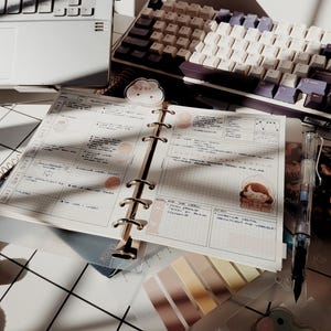 May include: An open ring-bound planner with grid paper, featuring handwritten notes, task lists, and decorative stickers of a character and a cloud. A clear fountain pen, a set of adhesive tabs, and a mechanical keyboard are visible nearby.