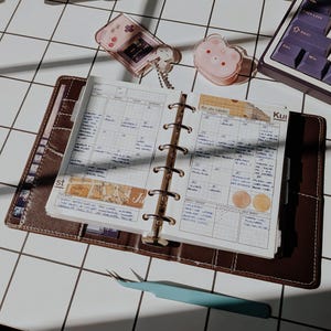 Peut inclure: Un agenda à six anneaux en cuir marron ouvert sur une page de calendrier mensuel avec des notes manuscrites, du papier quadrillé et du ruban adhésif décoratif. On y trouve aussi un petit porte-clés console rose, un charm rose et une pince bleue.