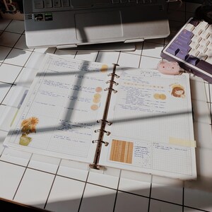 Peut inclure: Un classeur à six anneaux ouvert avec des pages à grille contenant des notes manuscrites, des autocollants circulaires jaunes, un autocollant de plante en pot, un personnage de dessin animé et un petit clip en forme d'ours sur une grille.