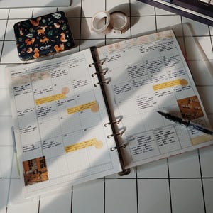 May include: A ring-bound planner open to a monthly calendar grid with handwritten notes, yellow sticky tabs, and decorative stickers. A black fountain pen rests on the page. Nearby sits a small rectangular tin with a forest animal pattern.