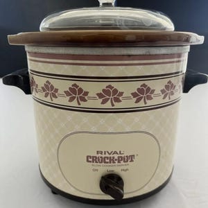 May include: A Rival Crock-Pot slow cooker with a cream-colored exterior featuring a brown diamond pattern and a band of brown leaf motifs. It includes a clear glass lid with a knob, black side handles, and a brown control dial labeled Off, Low, High.