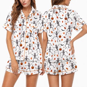 May include: A white pajama set featuring a repeating pattern of black skeletons holding orange jack-o'-lanterns, interspersed with orange autumn leaves, black bats, and small stars. The short-sleeved button-down top and shorts have orange piping.