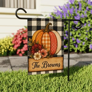 May include: A rectangular garden flag featuring a black and white buffalo check pattern, a large orange pumpkin with a plaid design, a cluster of red and white flowers, and a wooden sign with the black text The Browns hanging on a metal stand.