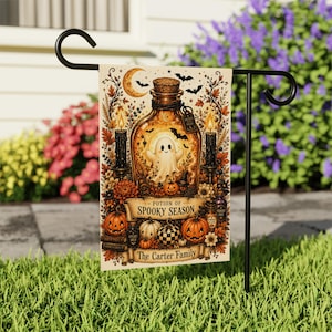 May include: A rectangular garden flag featuring a central glass bottle containing a ghost, surrounded by autumn leaves, pumpkins, candles, and bats. The text reads Potion of Spooky Season and The Carter Family on a cream-colored background.