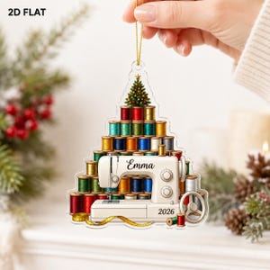 May include: A flat, tree-shaped acrylic ornament featuring a stack of colorful sewing thread spools topped with a small Christmas tree. A white sewing machine is centered at the base with the name Emma and the year 2026 printed on it.