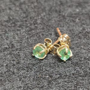 May include: A pair of stud earrings featuring round, faceted green gemstones set in gold-toned metal prongs. Each earring has a post back mechanism, showcasing a classic design with a vibrant green hue and a polished metallic finish on the setting.