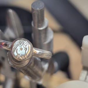 Könnte beinhalten: Ein silberfarbener Ring mit einer kreisförmigen Platte, die ein geprägtes Pferdekopf-Design aufweist. Der Ring wird von einem Metall-Ringstock gehalten, was das metallische Finish und das detaillierte Relief des Pferdeprofils deutlich zeigt.