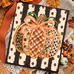 May include: A rectangular sign featuring a central pumpkin illustration with a peach-colored bow. The pumpkin has segments with leopard print, floral patterns, orange gingham, and polka dots. The background has vertical brown and white stripes.