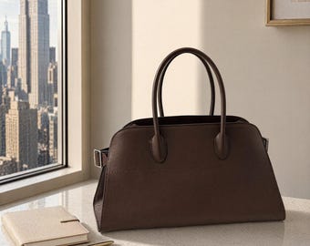 May include: A dark brown structured handbag with two top handles and a side buckle detail. The bag features a smooth, solid-colored exterior with clean lines and a wide base, positioned on a light-colored surface next to a small notebook and pen.