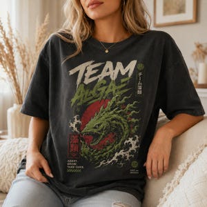 May include: A charcoal gray short-sleeve t-shirt featuring a graphic of a green, eel-like creature emerging from a red circle. The text reads TEAM ALGAE, ADAPT. GROW. TAKE OVER., and includes Japanese characters and small circular icons.