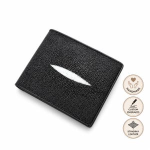 Può includere: Un portafoglio nero a due ante in pelle di razza testurizzata, caratterizzato da un inserto bianco allungato a forma di diamante sul davanti. A destra sono presenti tre icone circolari con le etichette HANDMADE, CUSTOM ENGRAVED e STINGRAY LEATHER.