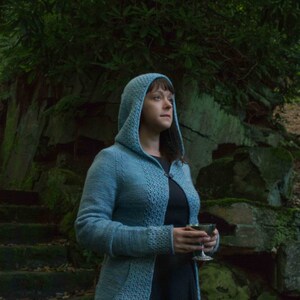 May include: A woman wearing a blue hooded cardigan with a lace pattern, standing in front of a mossy rock wall, holding a silver chalice.