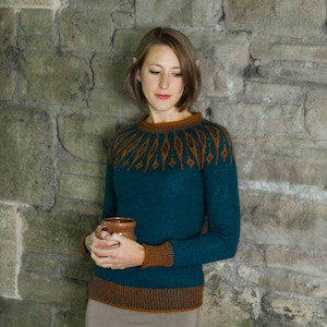 Könnte beinhalten: Eine Frau trägt einen tealblauen Pullover mit einem braunen und orangefarbenen geometrischen Muster. Der Pullover hat lange Ärmel und eine figurbetonte Silhouette. Sie hält eine braune Keramiktasse in ihren Händen.