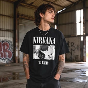 May include: A black short-sleeved t-shirt featuring the white text NIRVANA at the top, a black and white graphic image in the center, and the word BLEACH in white text below the graphic. The shirt has a crew neckline and a relaxed fit.