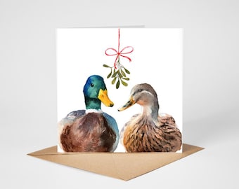 May include: A square greeting card featuring a watercolor illustration of a male mallard duck with a green head and a female mallard duck with brown feathers. A sprig of mistletoe hangs from a red ribbon bow centered above the two ducks.
