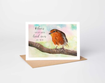 May include: A greeting card featuring a watercolor illustration of a robin perched on a brown tree branch. The text reads, Robins appear when loved ones are near. The background is a soft, pastel-colored wash of light blue, green, and pink hues.