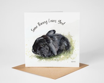 May include: A square greeting card featuring a watercolor illustration of a black lop-eared rabbit resting in green grass. The text Some Bunny Loves You! is printed in black script above the rabbit. A brown envelope is positioned underneath the card.