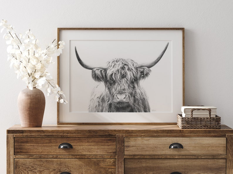 May include: Black and white pencil drawing of a Highland cow with long, shaggy fur and large horns. The drawing is framed in a simple wooden frame and is displayed on a wooden dresser.