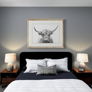 May include: A black upholstered bed with white and gray striped bedding and a throw pillow. Two bedside tables with lamps are on either side of the bed. A framed black and white print of a Highland cow is hanging on the wall above the bed.