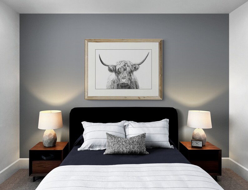 May include: A black upholstered bed with white and gray striped bedding and a throw pillow. Two bedside tables with lamps are on either side of the bed. A framed black and white print of a Highland cow is hanging on the wall above the bed.