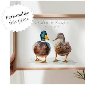 May include: A framed wall print featuring a watercolour illustration of two ducks standing side by side. The text reads JAMES & ALANA and 11. 05. 2024 with a small heart symbol. The male duck has a green head and yellow beak, while the female is brown.