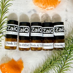 May include: Five small amber glass vials with black caps and white labels reading ZaiQ Fragrance. The labels identify the scents as Dusk Tide, Citrus Ether, Musk Al Tahara, Crimson Oud, and Smoky Vetiver. The vials are arranged horizontally.