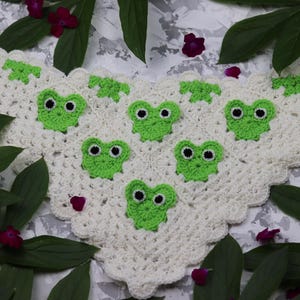 May include: A triangular crochet bandana in off-white yarn featuring a scalloped edge and six bright green frog motifs. Each frog has two black and white circular eyes. The piece is constructed with a repeating openwork stitch pattern throughout.