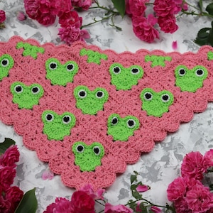 May include: A triangular crochet shawl featuring a pink yarn base with a scalloped edge. The shawl is decorated with multiple small, bright green crocheted frog motifs, each featuring two black and white circular eyes stitched onto the surface.