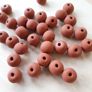 May include: A collection of terracotta-colored round beads with a hole in the center. The beads are scattered on a light wood surface.