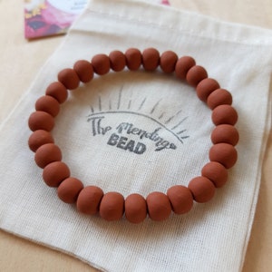 May include: A terracotta-colored beaded bracelet with a textured finish. The bracelet is displayed on a white fabric pouch with the text "The Mending Bead" printed on it.