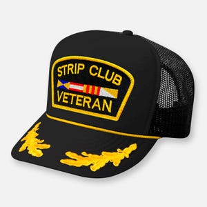 Puede incluir: Una gorra de camionero negra con un parche bordado en amarillo en la parte frontal que incluye el texto STRIP CLUB VETERAN y un gráfico de cinta. La visera presenta detalles de hojas bordadas en amarillo y un cordón amarillo.