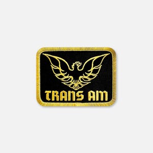 Puede incluir: Un parche rectangular bordado con fondo negro que presenta un emblema de águila estilizada en color dorado sobre el texto TRANS AM en una fuente gruesa y cuadrada, todo encerrado en un borde grueso cosido en hilo dorado.