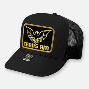May include: A black trucker hat featuring a rectangular patch on the front. The patch displays a gold embroidered eagle emblem above the text TRANS AM in gold lettering. A small black and white sticker on the brim reads OTTO WORLD FAMOUS FOAM FRONT.