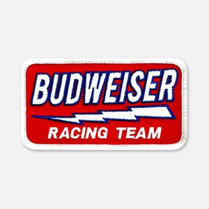 May include: A rectangular embroidered patch with a red background and white border. The text BUDWEISER is in white with a blue outline above a white lightning bolt graphic. The text RACING TEAM is in white letters at the bottom of the patch.