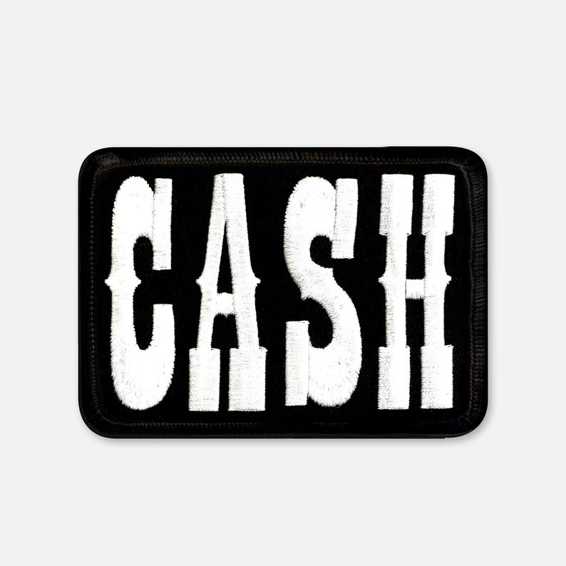 Pu&ograve; includere: Una toppa rettangolare ricamata con sfondo nero e la parola CASH cucita in grandi lettere maiuscole bianche e in grassetto. La toppa presenta un bordo nero spesso e cucito che circonda il testo bianco, creando un design ad alto contrasto.