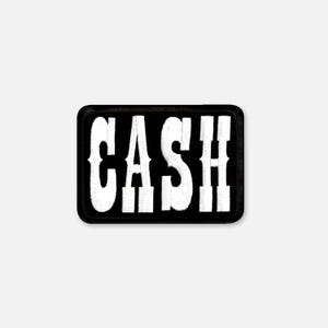 以下が含まれることがあります： 黒い背景に白い大きな太字のアルファベットでCASHと刺繍された長方形のワッペン。ワッペンには太い黒い縁取りがあり、全体的に糸のような質感のデザインが施されています。
