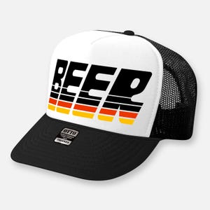 Könnte beinhalten: Eine weiß-schwarze Trucker-Cap mit dem Wort BEER in fetter, schräger schwarzer Schrift, unterlegt mit horizontalen roten und gelben Streifen. Die Kappe hat einen schwarzen Schirm, ein schwarzes Netzteil und einen Otto-Aufkleber.
