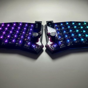 May include: A split ergonomic mechanical keyboard featuring a dark frame with individual key switches illuminated by purple and teal LED lights. The device includes a small vertical display screen and a circular rotary knob on each half of the unit.