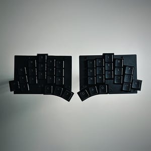 May include: A black split ergonomic keyboard featuring two separate rectangular modules with a staggered key layout. Each module contains black keys with white lettering, including letters, punctuation, and symbols, arranged in a non-traditional, contoured grid pattern for typing.