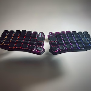 May include: A split ergonomic mechanical keyboard featuring two separate halves with black textured keycaps. The base is purple, and the keys are illuminated with a vibrant gradient of orange, pink, and blue light. A small digital display is visible.