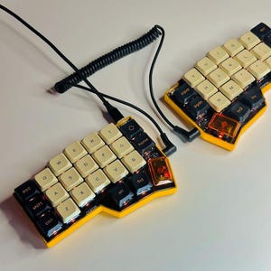 May include: A split ergonomic mechanical keyboard with a bright yellow chassis. The keycaps are a mix of cream and black, featuring standard QWERTY lettering. A black coiled cable connects the two halves, and each side includes a translucent orange key.