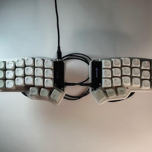 May include: A split ergonomic mechanical keyboard with two separate white keycap sections featuring black lettering. The halves are connected by a black coiled cable, with a central black bridge piece labeled Corne. The layout is ortholinear.