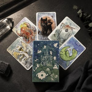 Peut inclure: Un jeu de cartes Lil Cryptid Oracle comprenant une boîte illustrée d'une forêt verte et cinq cartes individuelles présentant des dessins à l'aquarelle de créatures, dont une pieuvre, un loup, un homme-papillon et un serpent de mer avec texte.