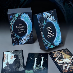 Puede incluir: La baraja de tarot The Lubanko Tarot y tres cartas individuales. La caja presenta ilustraciones en azul oscuro y verde azulado con el texto The Lubanko Tarot by E. Lubanko. Las cartas muestran dibujos detallados de una figura con una espada, dos pilares y figuras.