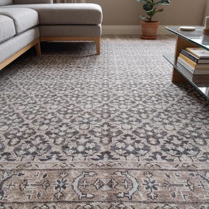 Könnte beinhalten: Ein rechteckiger Teppich mit einem sich wiederholenden floralen Medaillonmuster in Dunkelgrau und Beige. Der Teppich hat eine breite dekorative Bordüre mit komplexen geometrischen Motiven und eine kurze weiße Franse an der Unterkante.