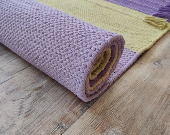 May include: A rectangular woven rug featuring alternating vertical stripes of pale yellow and light purple. The rug has a textured, knotted surface and is partially rolled up, revealing the inner layers. The edges are finished with short tassels.