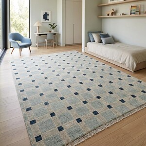K&ouml;nnte beinhalten: Ein rechteckiger Teppich mit einem Gittermuster aus hellblauen und beigen Quadraten, wobei sich an jedem Schnittpunkt ein dunkelblaues Quadrat befindet. Der Teppich hat eine strukturierte Oberfl&auml;che und kurze wei&szlig;e Fransen an den kurzen Seiten.
